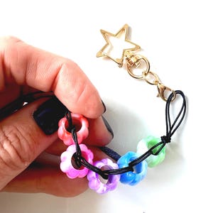 Può includere: Un portachiavi colorato con perline a forma di fiore nei toni del rosso, rosa, viola, blu e verde. Le perline sono infilate su un cordino nero e attaccate a una chiusura dorata a forma di stella.