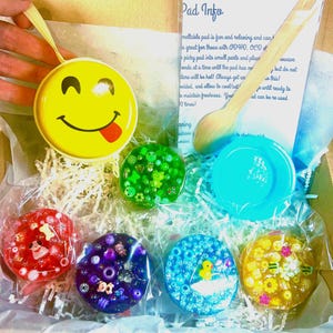 May include: A box containing a yellow smiley face container with a ribbon, a wooden spoon, and several colourful, round, meltable pads. The pads are in various colours and decorated with beads and small charms. The text "Pad Info" is visible.