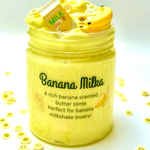 Może przedstawiać: Słoik żółtego masła "Banana Milka". Słoik wypełniony jest kremowym, żółtym slime'em i zwieńczony plasterkami banana oraz małym kartonikiem "Banana Milk". Na słoiku widnieje napis "A rich banana scented butter slime".