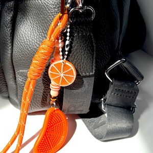 Op de afbeelding: Een zwarte tas met een oranje en witte bedel met een ontwerp van een citrusvrucht. De bedel is bevestigd aan een gevlochten oranje koord. Een klein oranje siliconen object is ook bevestigd. De tas heeft een textuur.