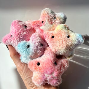 May include: A collection of five small, plush star-shaped toys with a soft, fuzzy texture. Each star features a tie-dye pattern in shades of pink, blue, yellow, and white, with two small, round, black plastic eyes embedded in the fabric.