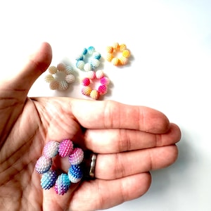 Può includere: Una collezione di anelli floreali colorati con perline. Gli anelli sono in vari colori, tra cui rosa, blu, giallo e bianco. Un anello è tenuto in una mano, mentre gli altri sono sparsi. Le perline hanno una superficie strutturata e irregolare.