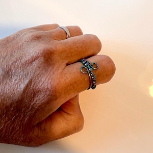 May include: An iridescent, multi-coloured metal ring featuring a series of small, round beads along the band and a decorative, looped wire design at the centre. The ring is worn on a finger, with a simple, plain silver-toned band visible on another finger.
