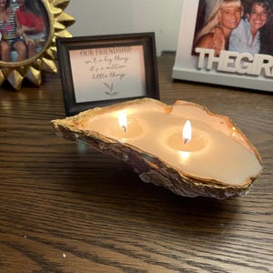 May include: A double-wick candle poured into a natural oyster shell with a gold-painted rim. The wax is off-white and illuminated by two flickering flames. The shell has a textured, irregular exterior and sits horizontally, showcasing the melted wax.