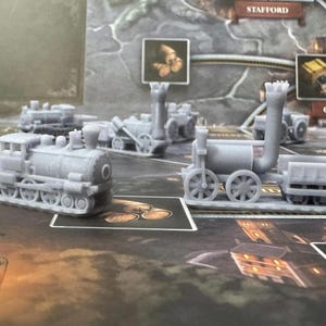 May include: Several gray miniature train models are arranged on a board game. The trains have detailed features, including wheels, smokestacks, and cargo cars. The game board features a map with place names.