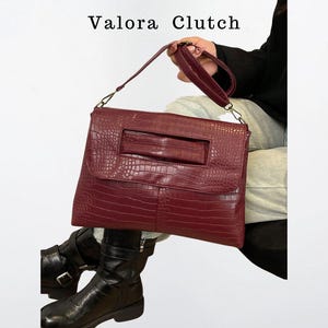 Peut inclure: Un sac &agrave; main Valora Clutch bordeaux avec une texture peau de crocodile. Le sac rectangulaire a une poign&eacute;e et une dragonne. Le sac est tenu par une personne portant des bottes noires.