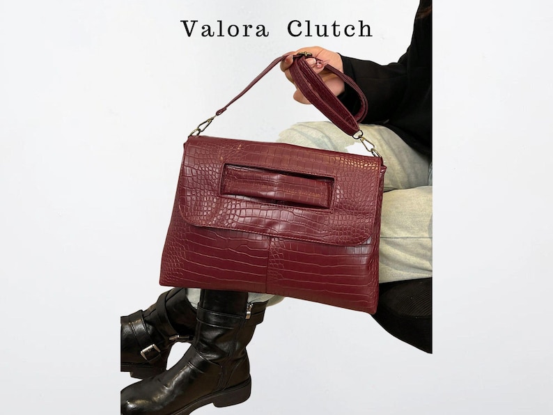 Peut inclure: Un sac &agrave; main Valora Clutch bordeaux avec une texture peau de crocodile. Le sac rectangulaire a une poign&eacute;e et une dragonne. Le sac est tenu par une personne portant des bottes noires.