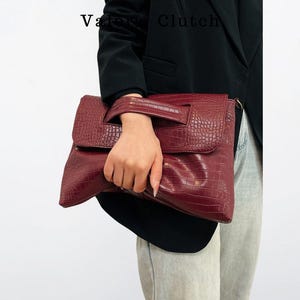 Peut inclure: Une pochette bordeaux avec une texture en peau de crocodile. La pochette a une fermeture &agrave; rabat et une poign&eacute;e. Les mots "Vatra Clutch" sont visibles dans le coin sup&eacute;rieur gauche.