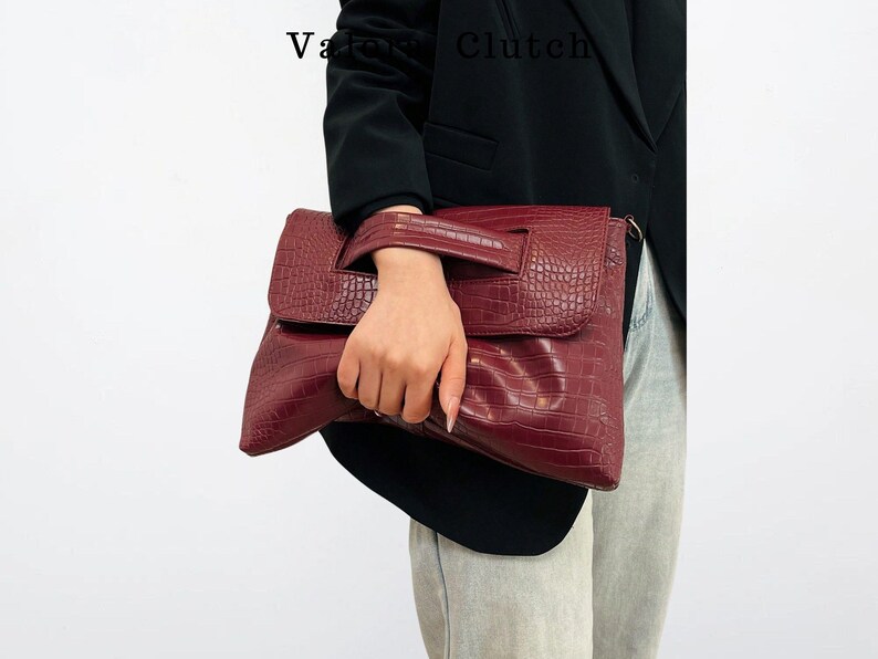 Peut inclure: Une pochette bordeaux avec une texture en peau de crocodile. La pochette a une fermeture &agrave; rabat et une poign&eacute;e. Les mots "Vatra Clutch" sont visibles dans le coin sup&eacute;rieur gauche.
