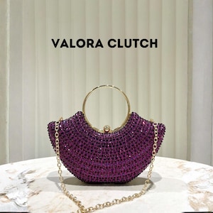 Puede incluir: Un bolso de mano morado con una correa de cadena dorada y un asa dorada. El bolso está cubierto de pequeñas gemas moradas brillantes. El texto "VALORA CLUTCH" se muestra arriba.