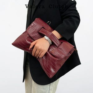 Peut inclure: Une pochette bordeaux avec une texture en peau de crocodile. La pochette a une poign&eacute;e et une fermeture &agrave; rabat. Le nom de la marque "Valora Clutch" est visible en haut &agrave; gauche. La pochette est tenue par une personne portant un blazer noir.