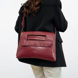 Peut inclure: Un sac &agrave; main bordeaux avec une texture en peau de crocodile. Le sac a une forme rectangulaire avec une fermeture &agrave; rabat et une poign&eacute;e. Le sac est port&eacute; &agrave; l'&eacute;paule avec une bandouli&egrave;re. Le texte "Valora Clutch" est visible.