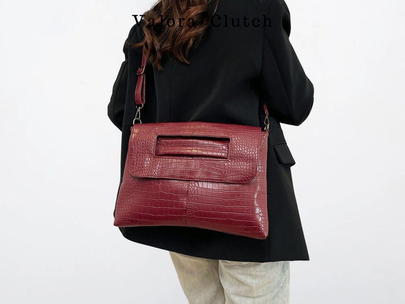 Peut inclure: Un sac &agrave; main bordeaux avec une texture en peau de crocodile. Le sac a une forme rectangulaire avec une fermeture &agrave; rabat et une poign&eacute;e. Le sac est port&eacute; &agrave; l'&eacute;paule avec une bandouli&egrave;re. Le texte "Valora Clutch" est visible.