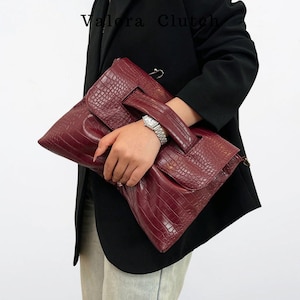 Peut inclure: Une pochette bordeaux avec une texture en peau de crocodile. La pochette a une poign&eacute;e et une fermeture &agrave; rabat. Le nom de la marque "Valora Clutch" est visible en haut &agrave; gauche. La pochette est tenue par une personne portant un blazer noir.