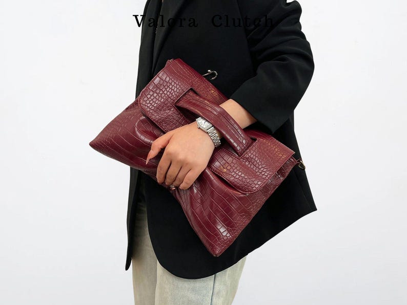 Peut inclure: Une pochette bordeaux avec une texture en peau de crocodile. La pochette a une poign&eacute;e et une fermeture &agrave; rabat. Le nom de la marque "Valora Clutch" est visible en haut &agrave; gauche. La pochette est tenue par une personne portant un blazer noir.