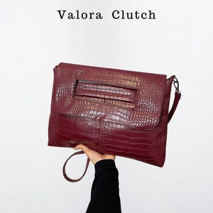 Peut inclure: Une pochette bordeaux avec une texture en peau de crocodile. La pochette est de forme rectangulaire avec un rabat et une poign&eacute;e. Le texte "Valora Clutch" est en haut de l'image.