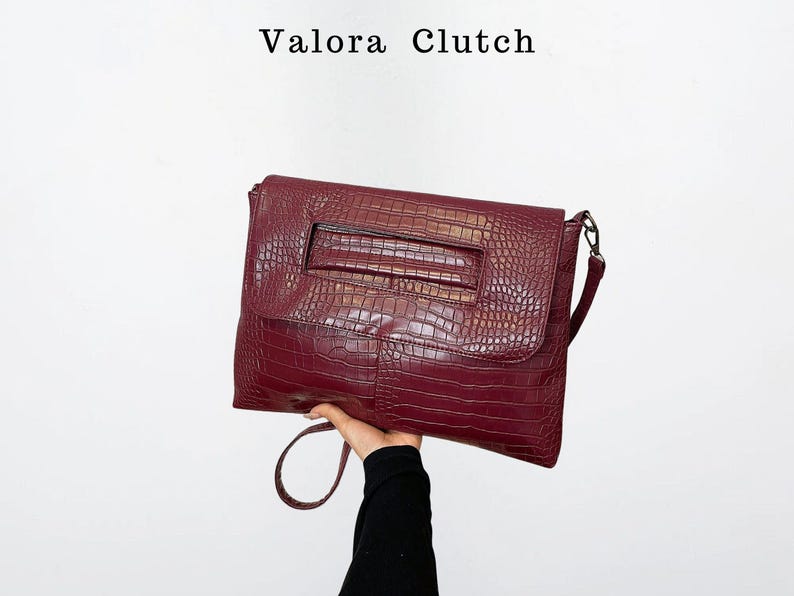 Peut inclure: Une pochette bordeaux avec une texture en peau de crocodile. La pochette est de forme rectangulaire avec un rabat et une poign&eacute;e. Le texte "Valora Clutch" est en haut de l'image.