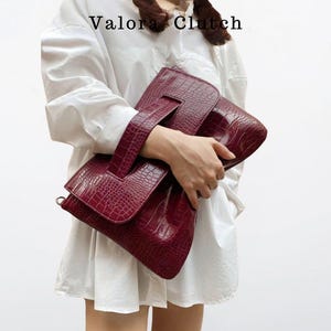Peut inclure: Une pochette bordeaux avec une texture peau de crocodile. La pochette a une poign&eacute;e et une fermeture &agrave; rabat. Le texte "Valora Clutch" est visible en haut. Le sac est tenu par une personne portant une chemise blanche.