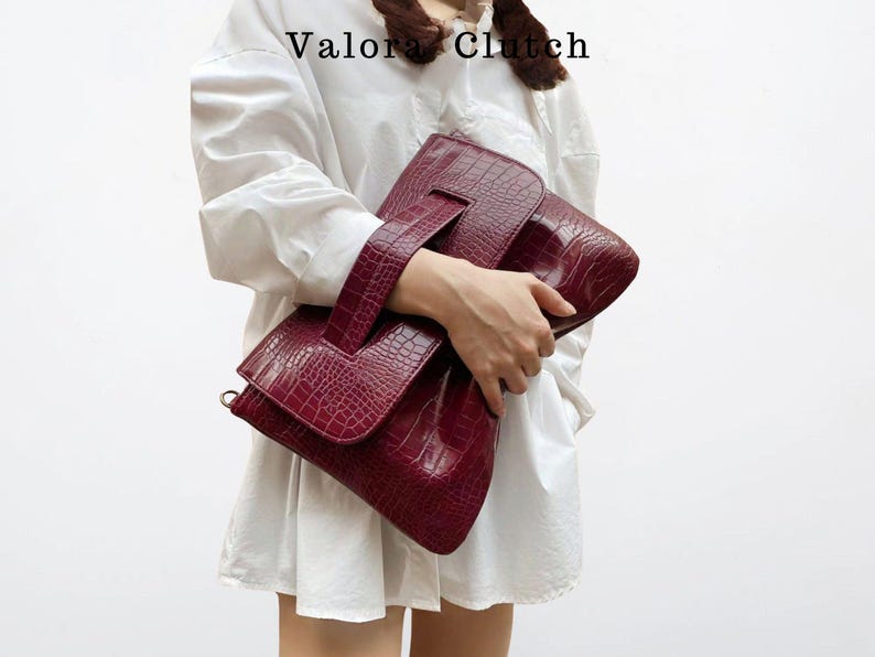 Peut inclure: Une pochette bordeaux avec une texture peau de crocodile. La pochette a une poign&eacute;e et une fermeture &agrave; rabat. Le texte "Valora Clutch" est visible en haut. Le sac est tenu par une personne portant une chemise blanche.