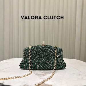 May include: A green beaded clutch with a gold chain strap. The clutch is rectangular with a rounded top and a small, decorative jewel clasp. The text "VALORA CLUTCH" is displayed above the clutch.