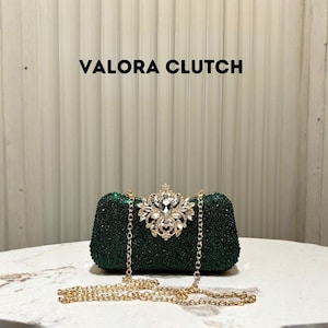 May include: A dark green clutch with a sparkling surface, featuring a decorative jeweled clasp. The clutch has a gold chain strap and the text "VALORA CLUTCH" is displayed above it.
