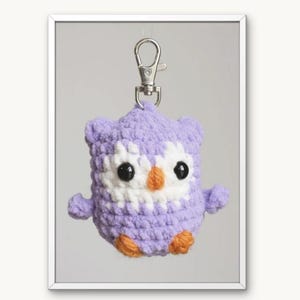 Può includere: Un portachiavi a forma di gufo lavorato all'uncinetto nei colori lavanda e bianco, con occhi in plastica nera e becco arancione. Il gufo è fissato a un moschettone in metallo argentato. Il testo POE THE OWL e The owl Amigurumi crochet pattern sono visibili.