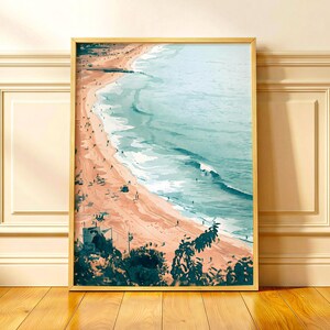 May include: A vertical framed wall art print featuring an aerial view of a sandy beach meeting the turquoise ocean. The scene includes small figures of people along the shoreline, a lifeguard tower in the foreground, and dark green foliage.