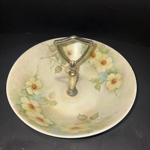 May include: A round porcelain serving dish featuring a hand-painted floral pattern of pale yellow flowers and green leaves. The dish includes a central metallic handle with a triangular top loop, finished in a polished silver-tone metal.