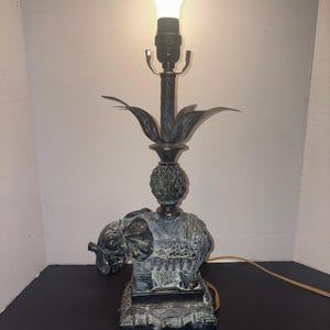 Puede incluir: Lámpara de mesa decorativa con base de elefante. La lámpara tiene un acabado oscuro y antiguo, un adorno en forma de piña y un diseño floral. La lámpara está rematada con una bombilla.