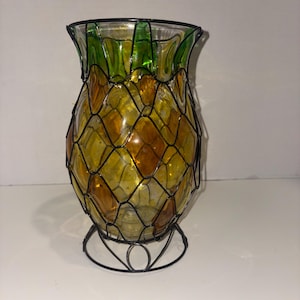 Puede incluir: Un jarrón decorativo de vidrio con forma de piña con una jaula y base de metal negro. El jarrón presenta paneles de vidrio en forma de diamante amarillos y naranjas, rematados con hojas de vidrio verdes. La base es un diseño de metal circular.