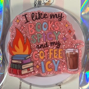 May include: A pink glittery phone grip with the text "I like my BOOKS SPICY and my COFFEE ICY." The design includes a stack of books with flames and a coffee drink with a straw. The phone grip is round and has a clear base.