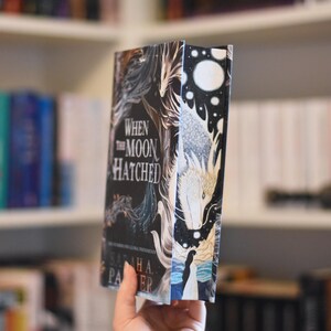 Könnte beinhalten: Ein Hardcover-Buch mit dem Titel "When the Moon Hatched" mit einem Fantasy-Cover. Das Cover hat einen dunklen Hintergrund mit Illustrationen eines Drachen, einer Frau und himmlischen Elementen. Der Buchrücken ist weiß mit schwarzen Punkten.
