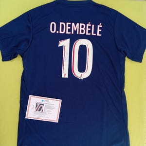 May include: A navy blue sports jersey featuring the name "O. DEMBÉLÉ" and the number "10" in white. The jersey has a signature on the number. A certificate of authenticity is visible.