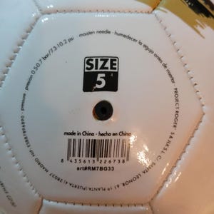 May include: A white soccer ball featuring a black valve and printed text including SIZE 5, pressure 0.5-0.7 bar/7.3-10.2 psi, moisten needle, made in China, art#RM7BG33, and a barcode. The surface has stitched hexagonal panels and gold accents.