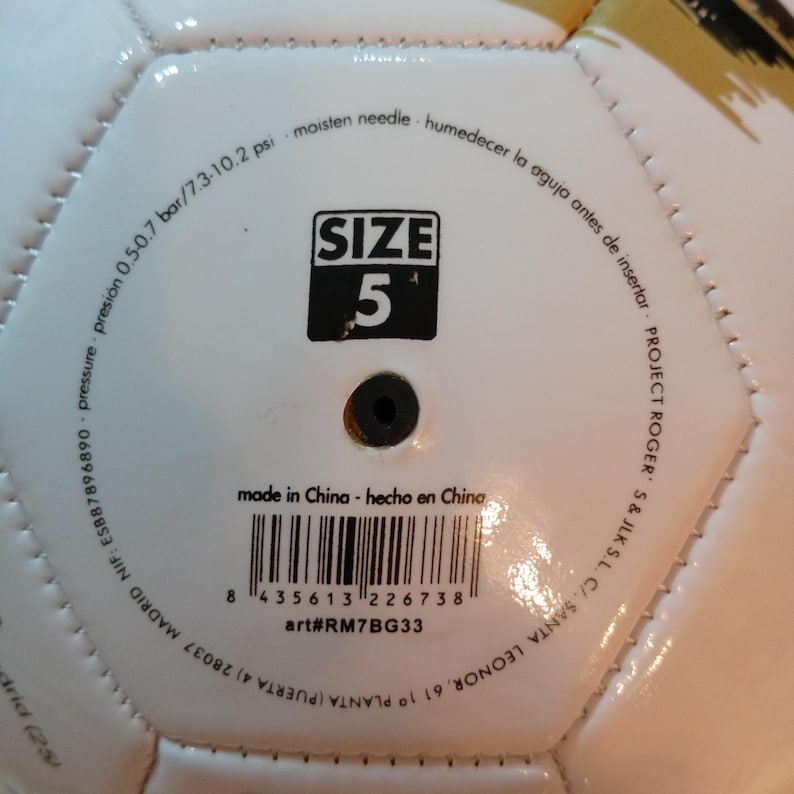 May include: A white soccer ball featuring a black valve and printed text including SIZE 5, pressure 0.5-0.7 bar/7.3-10.2 psi, moisten needle, made in China, art#RM7BG33, and a barcode. The surface has stitched hexagonal panels and gold accents.