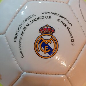 May include: A white soccer ball featuring the Real Madrid C.F. logo in blue, gold, and red. The text reads PRODUCTO OFICIAL, Con licencia del REAL MADRID C.F., www.realmadrid.com, and Real Madrid (25) printed in black around the central crest.
