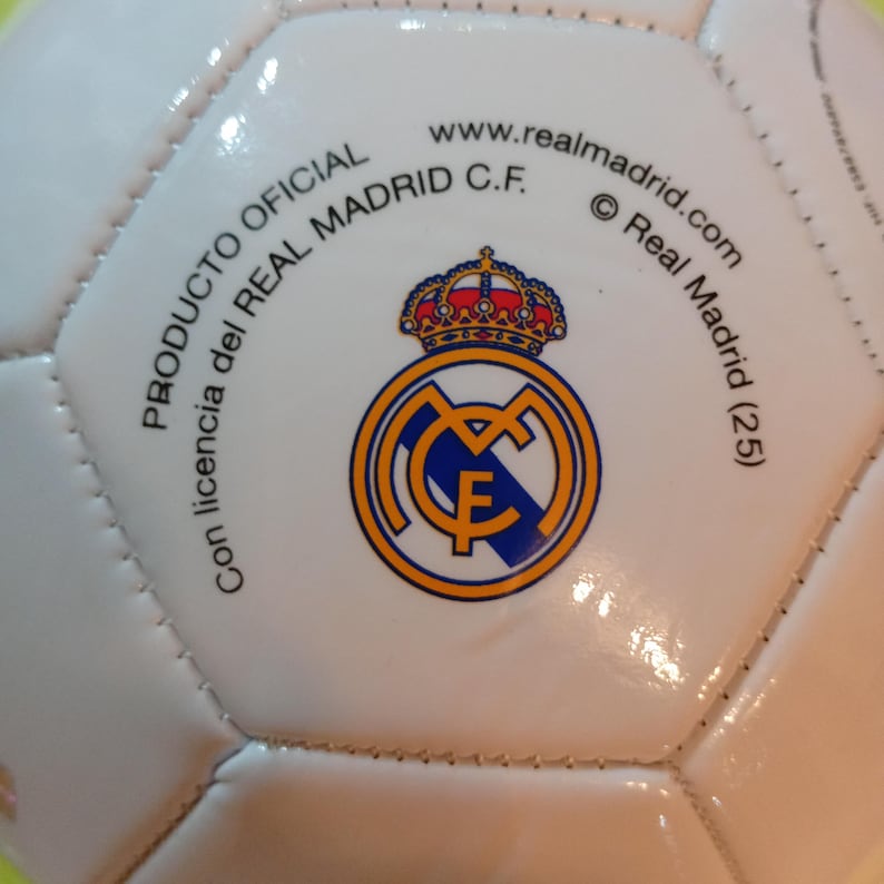 May include: A white soccer ball featuring the Real Madrid C.F. logo in blue, gold, and red. The text reads PRODUCTO OFICIAL, Con licencia del REAL MADRID C.F., www.realmadrid.com, and Real Madrid (25) printed in black around the central crest.