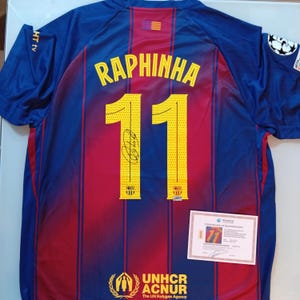 May include: A blue, red, and yellow soccer jersey with the name "RAPHINHA" and the number "11" on the back. The jersey has a signature and a certificate of authenticity. The UNHCR ACNUR logo is visible.