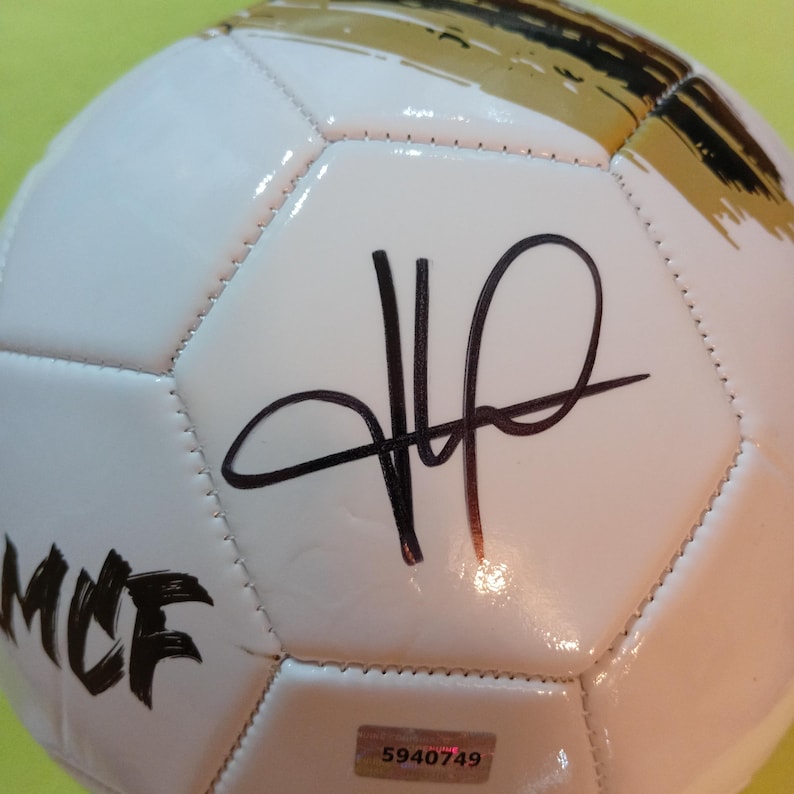 May include: A white soccer ball featuring a black signature in the center, the letters MCF in black on the side, and a gold graphic accent at the top. A small rectangular authentication sticker with the number 5940749 is affixed to the surface.