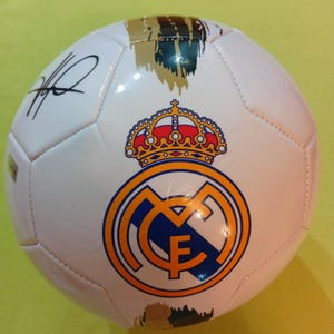 May include: A white soccer ball featuring the Real Madrid club crest in blue, gold, and red with a crown on top. The ball includes black brushstroke accents and a black ink signature on the upper left panel, along with a small rectangular label.