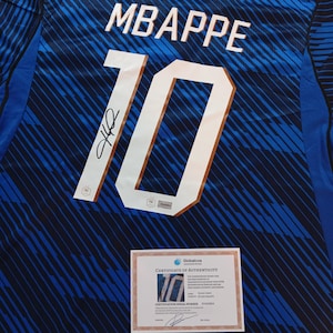 Puede incluir: Una camiseta de fútbol azul con un patrón abstracto azul oscuro, con el nombre MBAPPE y el número 10 en blanco. Una firma negra aparece sobre el número 10. Se incluye un certificado de autenticidad de Globalcoa con la camiseta.