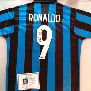 May include: A short-sleeved sports jersey featuring vertical black and blue stripes. The back displays the name RONALDO in white letters above a large white number 9 with a black signature. A certificate of authenticity is placed at the bottom.