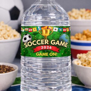 May include: Clear plastic water bottle with a white cap. The label features a soccer ball, a gold trophy, and the words "Soccer Game 2026" with "Game On!" in red. The background includes green grass and international flags.