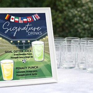 May include: A white framed sign with "Signature Drinks" text, featuring two drink options: Goal Getter Juice and Penalty Punch. The sign has a soccer field background and a string of flags. Clear glasses are arranged on a white surface.