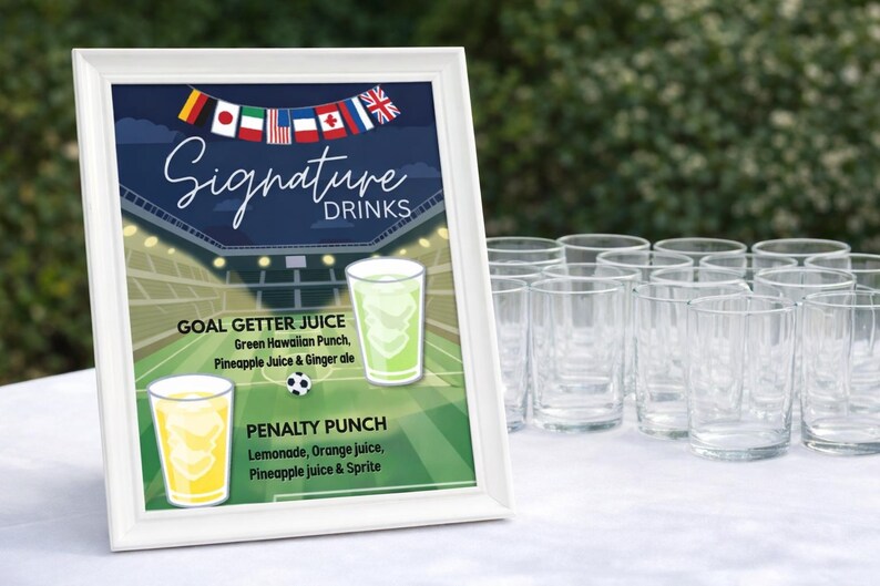 May include: A white framed sign with "Signature Drinks" text, featuring two drink options: Goal Getter Juice and Penalty Punch. The sign has a soccer field background and a string of flags. Clear glasses are arranged on a white surface.