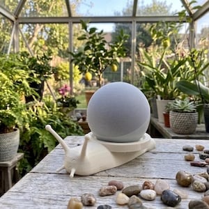 Peut inclure: Un support en forme d'escargot imprimé en 3D blanc tenant une enceinte connectée sphérique gris clair. L'escargot présente deux longues antennes et un visage souriant gravé sur le devant. L'appareil repose sur la plateforme circulaire plate.