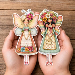 Pode incluir: Dois adesivos recortados com personagens de fadas ilustrados. A fada da esquerda tem cabelo loiro, asas cor-de-rosa e um vestido com detalhes florais. A fada da direita tem cabelo escuro, um grande chapéu de flores e uma pequena bolsa.