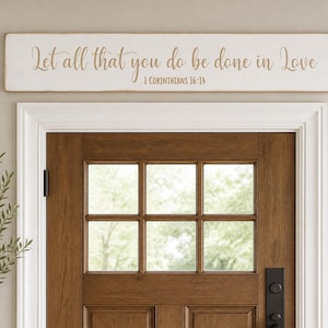 May include: A rectangular white wooden sign with distressed edges featuring the brown script text Let all that you do be done in Love and the citation 1 Corinthians 16:14, mounted horizontally above a brown wooden door with glass panes.