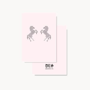 May include: Two pale pink rectangular cards. The top card features two mirrored rearing horses composed of small black dots. The bottom card displays the black text DES ARLENE CO. in a gothic-style font centered below the horse graphic.