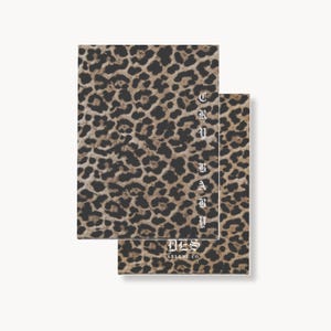 May include: Two rectangular notebooks featuring a brown and black leopard print pattern. The front notebook displays the vertical white text CRY BABY and the horizontal text DES ARLENE CO. in a gothic-style font on the lower portion.